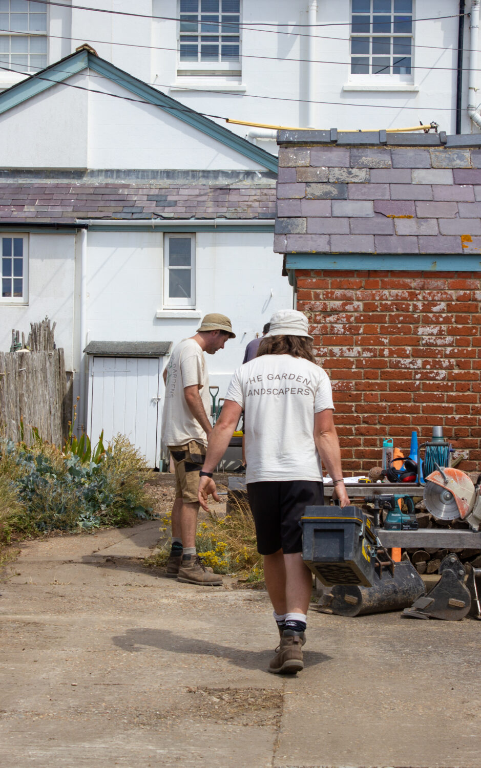 2 Garden landscapers one carrying a toolbox working on site in concreted front garden at the seaside.