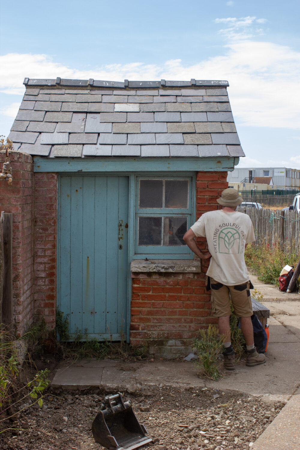 A Garden Landscaper facing away wearing tshirt printing with "Creating Soulful Gardens" around an arch shaped logo in a cosy beach garden in front of small red brick building with sky blue paintwork.